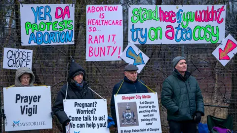 PA Media Pro-life protesters stand in zipped up coats and hats and hoods holding signs urging women to rethink abortion choices. Neon hand-made pro-choice signs in counter protest are hung on a fence behind them.