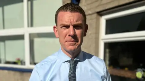 BBC A man is looking directly into the camera. He is wearing a light blue shirt and a dark patterned tie. The background is out-of-focus, but you can make out the wall of a school with a run of windows and flowerbeds. 