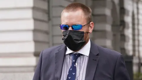 PA Media PC Craig Carter wearing a suit, tie, face mask and sunglasses