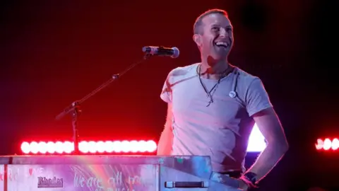 Reuters Coldplay lead singer Chris Martin on stage. He is wearing an off-white t-shirt and sitting behind a piano and microphone. A strip of red lights are visible behind him. 