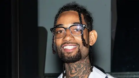 Getty Images Rapper PnB Rock, in New York in 2020, wearing glasses with a black rim. Smiling. He is wearing a white top, and has tattoos on his neck. The background is of a window.