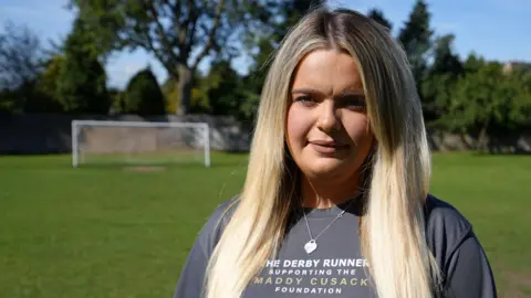 BBC Olivia Cusack pictured at a football field in Derby on a sunny day. Olivia has long blonde hair worn loose and wears a grey T-shirt promoting the Maddy Cusack Foundation. Behind her is a line of trees and a goal. 