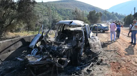 Getty Images A damaged burnt out car on the outskirts of Masyaf lies on the side of a dirty road in Syria. People watch on and take photos of the damaged car.