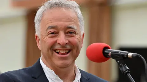 BBC A smiling Frank Cottrell-Boyce, with short grey hair and wearing a dark jacket and white shirt, stands in front of a red microphone