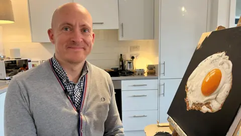 BBC/Corinne Wheatley Daniel Halksworth in a grey sweater and chequered shirt. He is smiling at the camera as he sits in his kitchen next to one of his fried egg paintings which is popped up on an easel on a table.