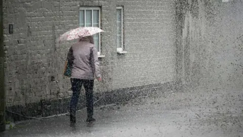 Getty Images A woman walking away from the camera in heavy rain. She is wearing a grey jacket, with a bag over her left shoulder. She is carrying a floral umbrella. 