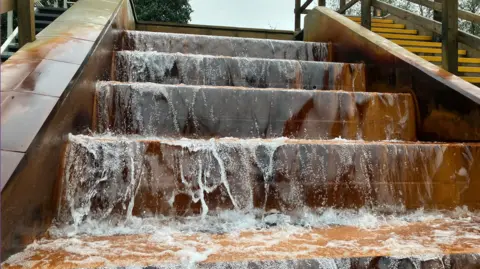 BBC Water which has been pumped from a mine shaft is cascading down four large steps which have turned an ochre colour