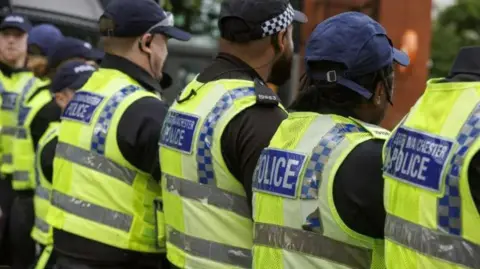 PA Media Five police officers stand with their backs to the camera, wearing hi-vis jackets with the blue badges on the back that reads Greater Manchester Police or just the word, Police. 