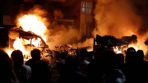 Instagram/Reuters Vehicles burning after being set alight in Harehills, Leeds, in July. Pictured are the flames and smoke billowing in the dark, while shadowed figures stand with their backs to the camera