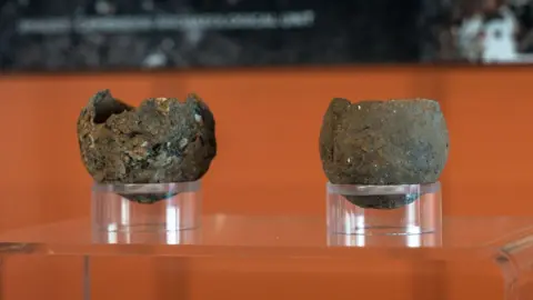 Martin Rowe/Peterborough Museum and Art Gallery Close up views of two Bronze Age pinch pots displayed in an exhibition case against an orange background at Peterborough Museum