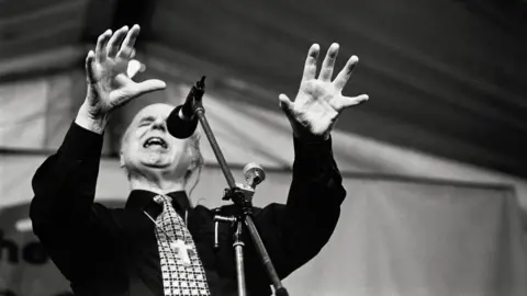 John Angerson Black and white image of the the leader of the Jesus Fellowship, Noel Stanton, giving a sermon with his arms raised in front of him