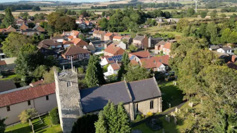 BBC A church and several houses in Shiptonthorpe can be seen from above. The sun has cast a shadow on the church and highlights different hues of green within nearby trees.