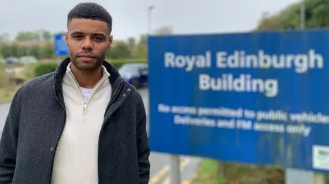 BBC Christopher MacRae, wearing a cream fleece and grey coat, standing in front of a sign for the Royal Edinburgh Hospital