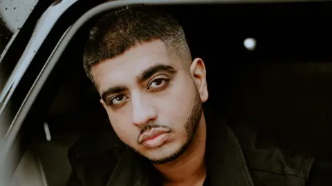 Jeff Rogers A man sitting in a car, looking outside with a serious expression. he is wearing a black jacket.