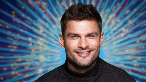 BBC Pictures/Nicky Johnston/PA Wire Aljaz Skorjanec faces the camera wearing a black turtle neck jumper, smiling. He has spiky dark hair. Behind him is a blue starburst image