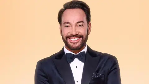 PA Media Craig Revel Horwood smiling dressed in a black tuxedo with his arms folded