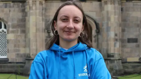 Aberdeen University Students' Association Christina Schmid, a woman with long brown hair, a blue hoodie and nose piercings, looks into the camera