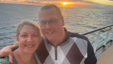 BBC Megan and Michael are standing on a boat with a sunset over the sea behind them. Michael has his arm around Megan's shoulder and they are smiling at the camera