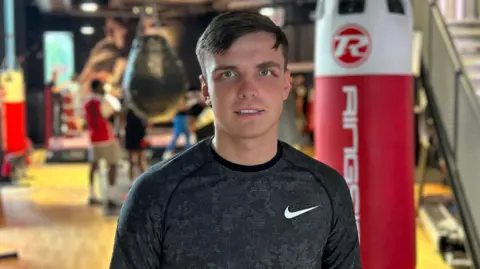 BBC Tommy Murphy, a 23-year-old man with short brown hair and blue eyes, pictured wearing a Nike jumper standing in front of a boxing gym.