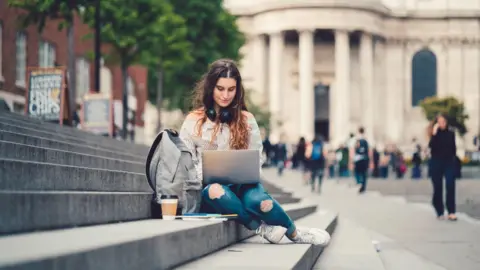 Getty Images A girl wearing blue jeans and a stripy black and white shirt is sitting on concrete steps on her laptop. She has a bag and a coffee beside her. She is outside an old grey building with people walking past. 