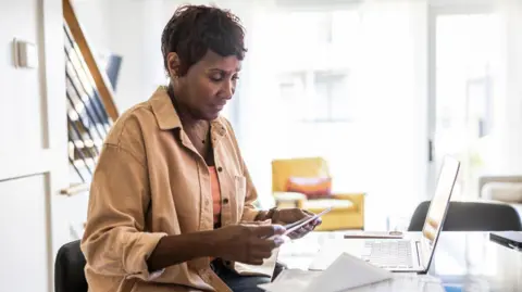 Getty Images A woman looks at a an envelope with bills while sitting in front of a laptop