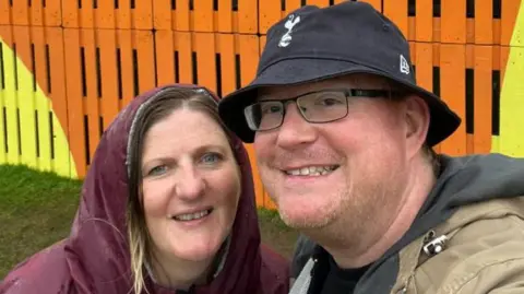 PA Media Kate and Colin Broadbent are smiling at the camera in front of orange and yellow wood panels. Colin is wearing a black bucket hat and black glasses, and Kate has brown hair, and is wearing a burgundy raincoat with the hood up