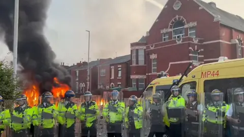 PA Media Line of riot police in high vis clothing holding riot shields outside mosque where one police van is on fire 