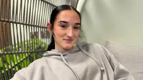 A young woman with dark hair in a ponytail, and an eyebrow piercing, wearing a light grey hoodie smiles while looking directly at the camera