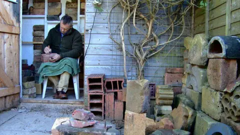 Mark Cranston sitting on a white plastic chair writing on a brick. He has a green blanket on his knees and is wearing a dark fleece. He is sitting in the doorway of his stables.