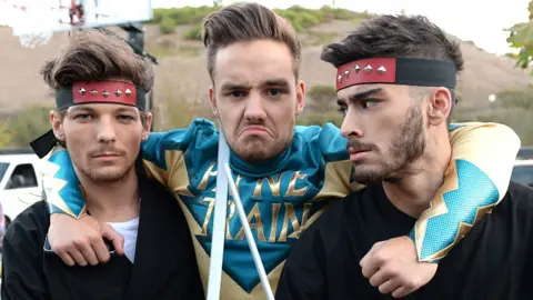 Getty Images Liam Payne posing and pulling a face in a superhero "Payne Train" costume, with his arms around the necks of Louis Tomlinson and Zayne Malik, who are wearing superhero-style headbands