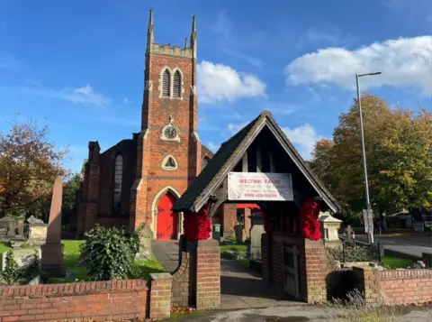 A church with orange bricks and a red archway, surrounded by graves and a brick wall. There are trees to the right and one on the left.