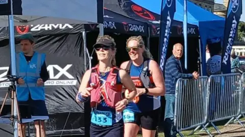 Contributed Mrs Hayward-Brackenbury is pictured running a race. She is running with a friend who is slightly behind her. Mrs Hayward-Brackenbury  is wearing a blue vest with a number on it, with a red vest over the top that carries water. She is wearing a black visor cap and black sunglasses.