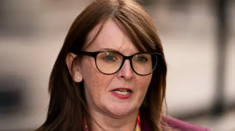 PA Media Caoimhe Archibald with brown/copper long hair and a side fringe. She is wearing dark framed glasses, a diamond stud earring in the ear not covered by her hair. She is mid-speech outside the Treasury in London. She is wearing a pink jacket and pink, red, yellow and orange floral scarf. 