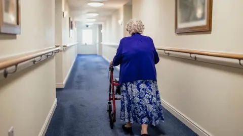 Getty Images An elderly woman uses a walker to go down an empty corridor with hand rails at either side and pictures on the wall.