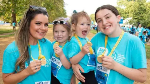 Louise Hubbard Louise Hubbard with her children Florence, Fleur and Felicity. They are all standing in a park wearing blue T-shirts, holding medals and smiling at the camera.