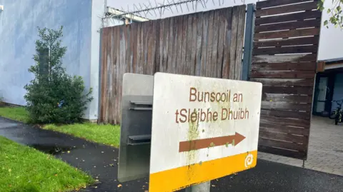 A white, yellow and brown sign pointing to the entrance of Bunscoil an tSléibhe Dhuibh.  It is located in front of a wooden barrier with anti-climb security spikes on the top of the wood.