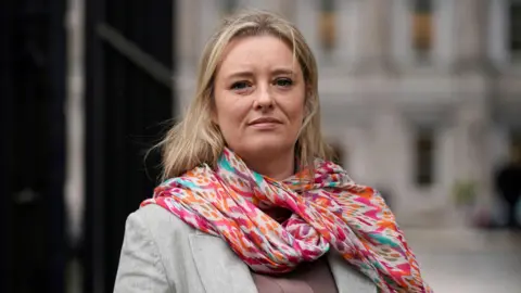 PA Images Máiría Cahill stands in front of the camera. She has blonde hair, a grey jacket and a colourful scarf. Her expression is neutral.