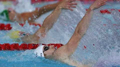 Getty Images a swimmer doing the backstroke, his white cap and black goggles peak out from the water, his arm is extended and surrounding by water splashes.