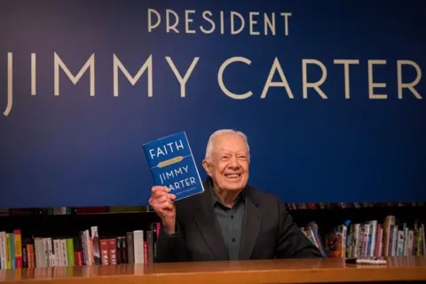 Getty Images Carter holds up a copy of his book Faith: A Journey For All at a book signing event in New York in 2018