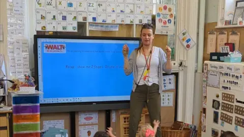 Amy Anderson Amy stood at the front of a class in front of a board and teaching children