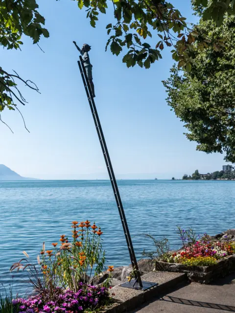 Sean Knight Sculpture of figure reaching out from the top of a ladder next to a lake