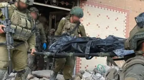 IDF Israeli soldiers with their faced blurred carry a corpse in a black body bag, purportedly of Yahya Sinwar, out of a destroyed house which has distinctive tilework on the front. 