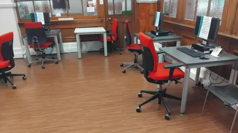 Contributed A room in an Edinburgh library in which all computers are turned off, with notices on the screen detailing they cannot be used due to a 'technical fault'. The office chairs pulled up the at the desks on the dark wooden floor are red, with a black back and frame.