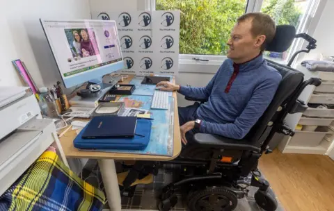 My Name'5 Doddie Foundation Scott Stewart sits in his wheelchair in his house in Stirling facing his computer where he works to earn a living for his family 