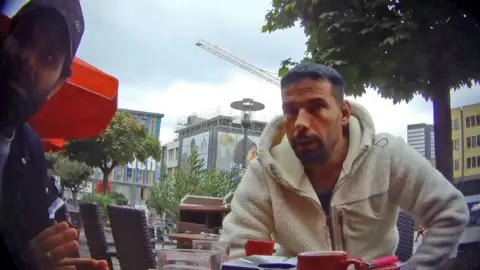 The dark, short-haired man with a beard and short hair wearing a white fleece jacket from previous photos captured by the undercover reporter engaged in discussion at a pavement cafe in central Essen. There are coffee cups on the table, high-rise buildings, trees and a crane in the background. A second man - with the Gucci baseball cap and blue shirt - with beard and dark hair can be seen on the far left of the shot. 