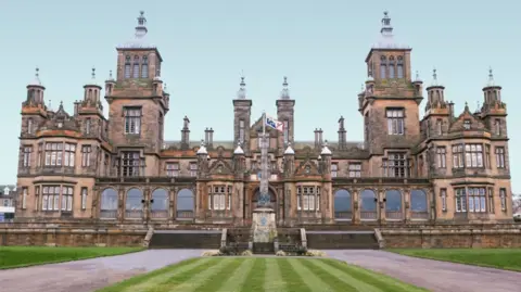 Google A large historic school building with mutliple towers and turrets, high arched windows, with a flagpole front and centre behind a manicured striped green lawn.