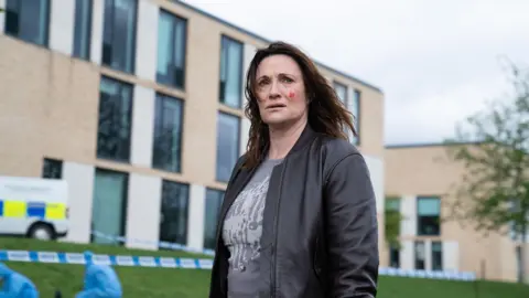 Curfew / Paramount+  Sarah Parish stands outside a tall building, there is police tape, a police van and team in the shot. She has blood on her cheek and stares into the distance, looking worried.