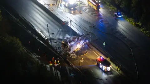 Eddie Mitchell An aerial shot of a burnt out vehicle on a section of motorway. Fire hoses are visible.