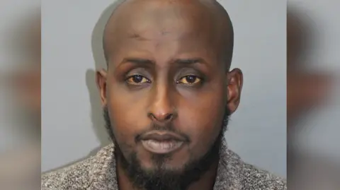 Met Police A close up police shot of Mohamed. He is bald with facial hair. Standing against a grey wall. 