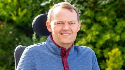 My Name'5 Doddie Foundation Scott Stewart smiles at the camera while sitting outside in his wheelchair in the sunlight with bushes in the background